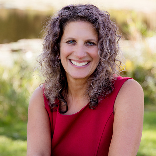 Photo of Sandy Wolf, therapist for couples and families. She is a white woman with shoulder-length curly brown hair wearing a red shirt. She is smiling at the camera and is outdoors on a sunny day with green grass and trees in the background.